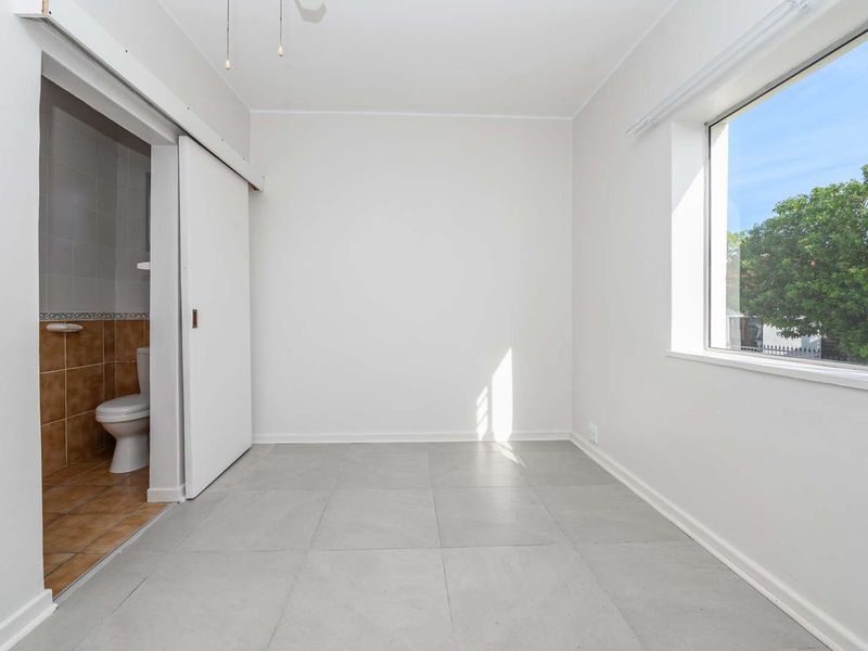 A bright, empty bedroom featuring neutral tiled flooring, a large window, and direct access to an en-suite bathroom via a sliding door.