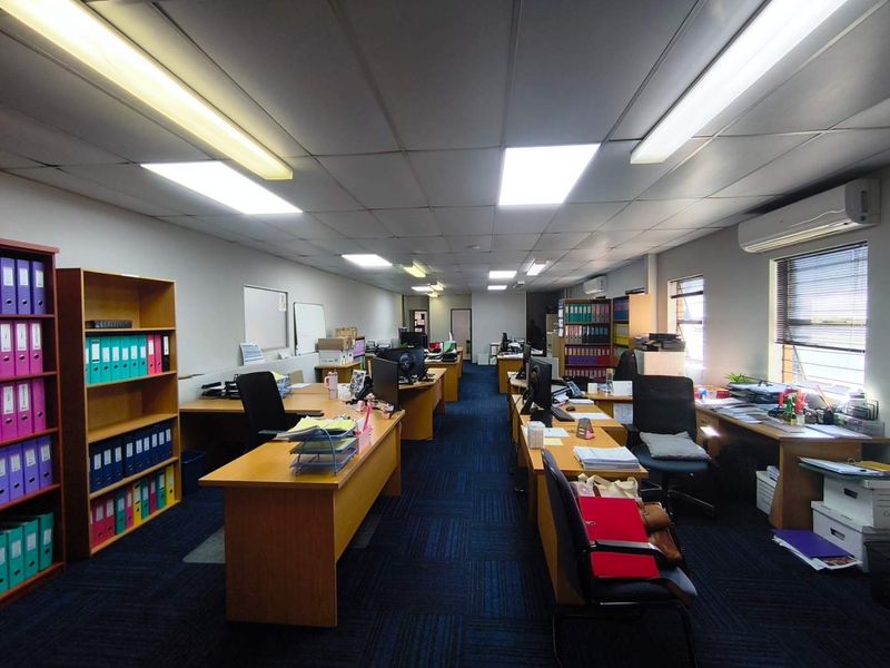 A functional open-plan office space featuring suspended ceilings, fluorescent lighting, carpet tiles, and wall-mounted air conditioning units.