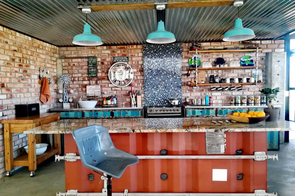 Industrial-style kitchen featuring a unique shipping container island, granite countertop, exposed brick walls, and corrugated metal ceiling with pendant lighting.