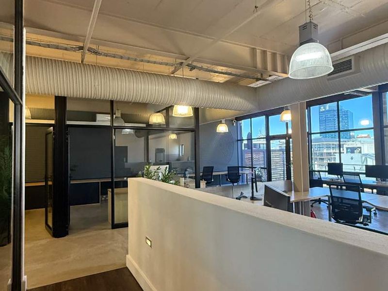 Modern open-plan office space featuring industrial-style exposed ceilings, ductwork, glass partitions, and large windows providing ample natural light and city views.