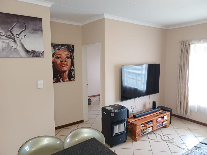 A living room area featuring tiled flooring, neutral wall tones, and a wall-mounted television setup with access to an adjacent hallway.