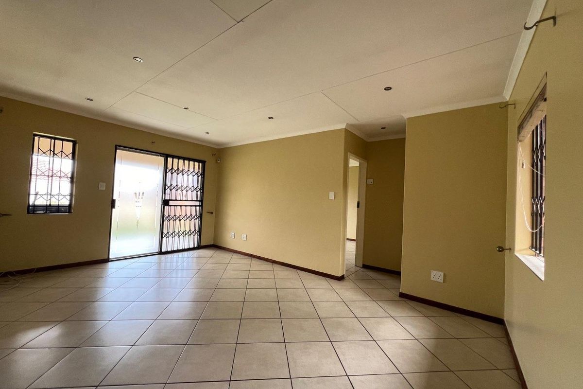A spacious, empty living area featuring neutral tiled flooring, recessed lighting, and secure access via a sliding glass door with security gate.