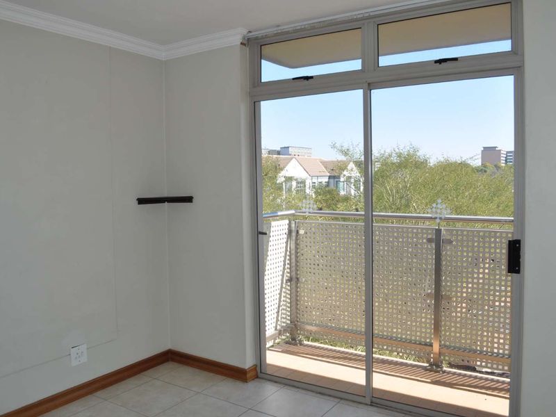 A bright, empty bedroom featuring tiled flooring, crown molding, and a sliding glass door that opens onto a private balcony with neighborhood views.