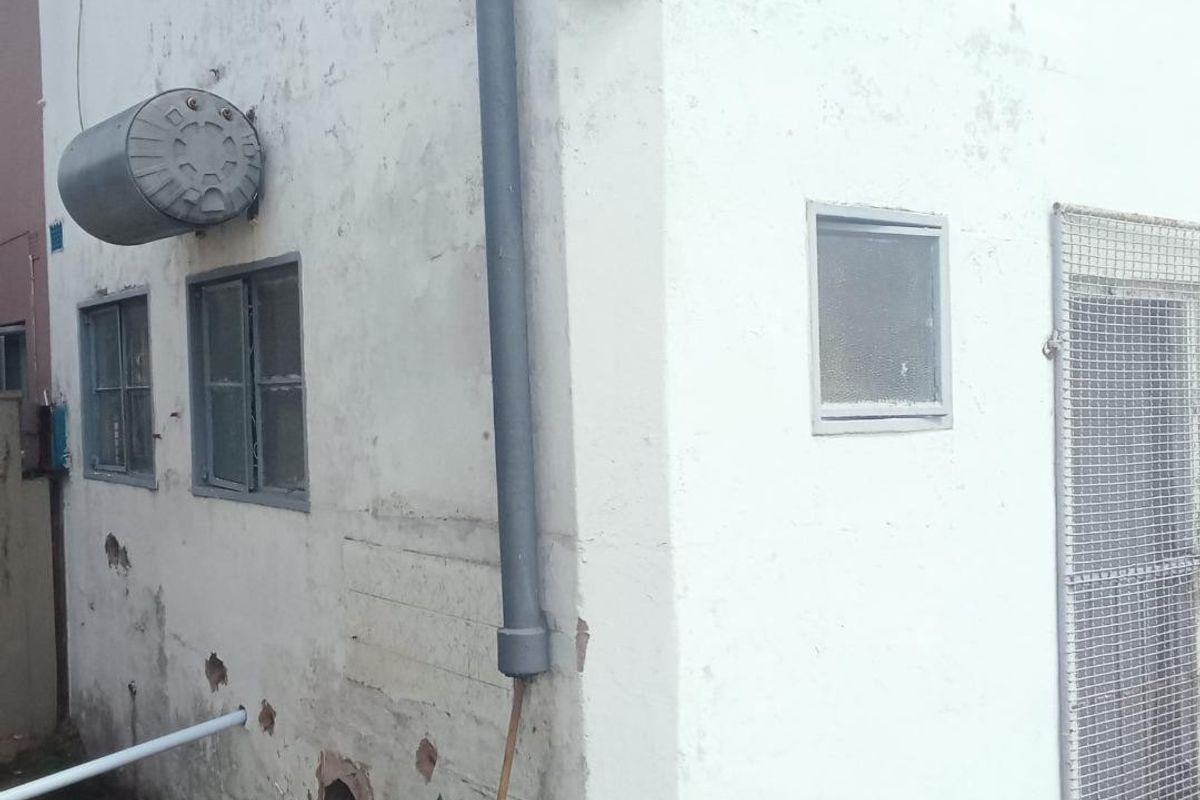 Rear exterior view of the property showing weathered wall finishes, an external geyser, and security features on the windows and door.