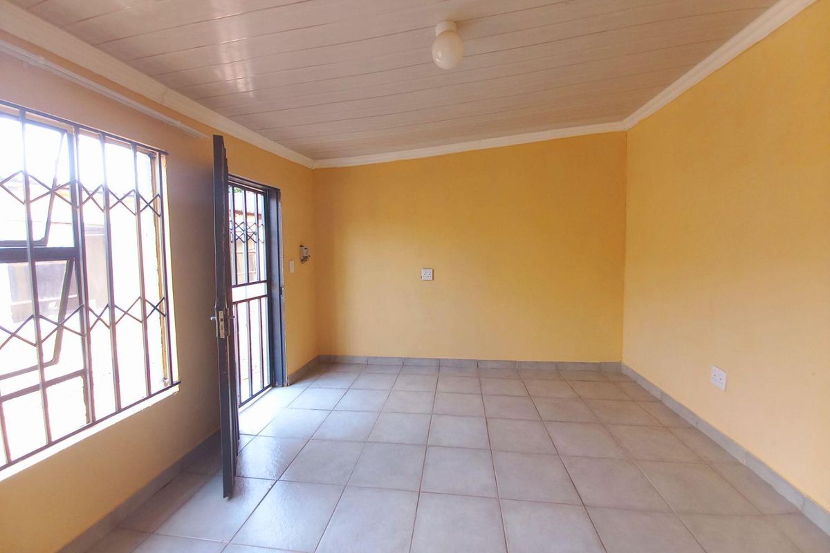 A bright, empty living room featuring tiled flooring, a white panelled ceiling, and security bars on the windows and door.