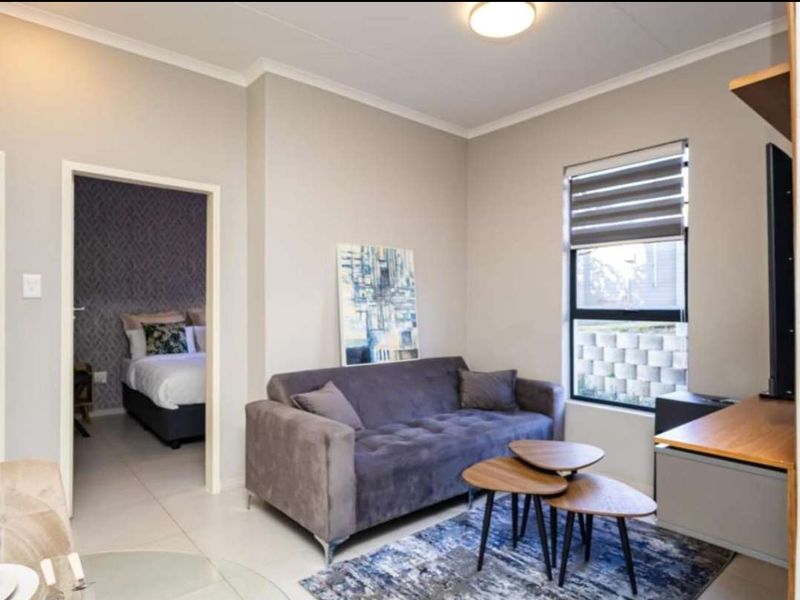 A well-maintained living room featuring neutral wall tones, tiled flooring, and a window with fitted roller blinds, leading into an adjacent bedroom.