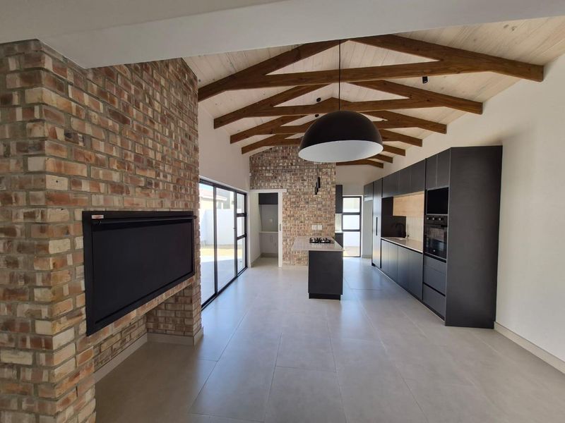 Spacious open-plan living and kitchen area featuring exposed brickwork, vaulted timber ceilings, modern dark cabinetry, and a built-in fireplace.