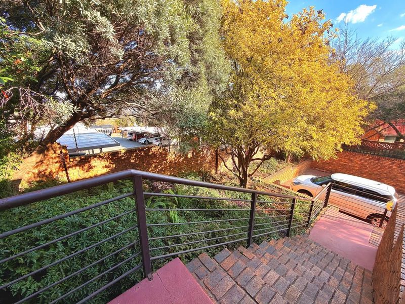 A view looking down a brick staircase with a metal handrail, leading into a landscaped garden area with mature trees and a brick boundary wall.