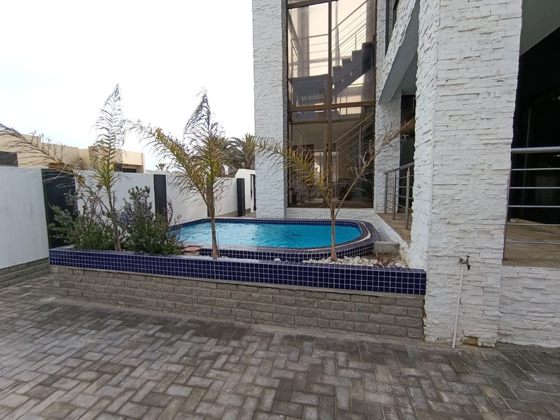 A modern outdoor area featuring a raised swimming pool with blue tile finish, stone-clad exterior walls, and paved patio space.