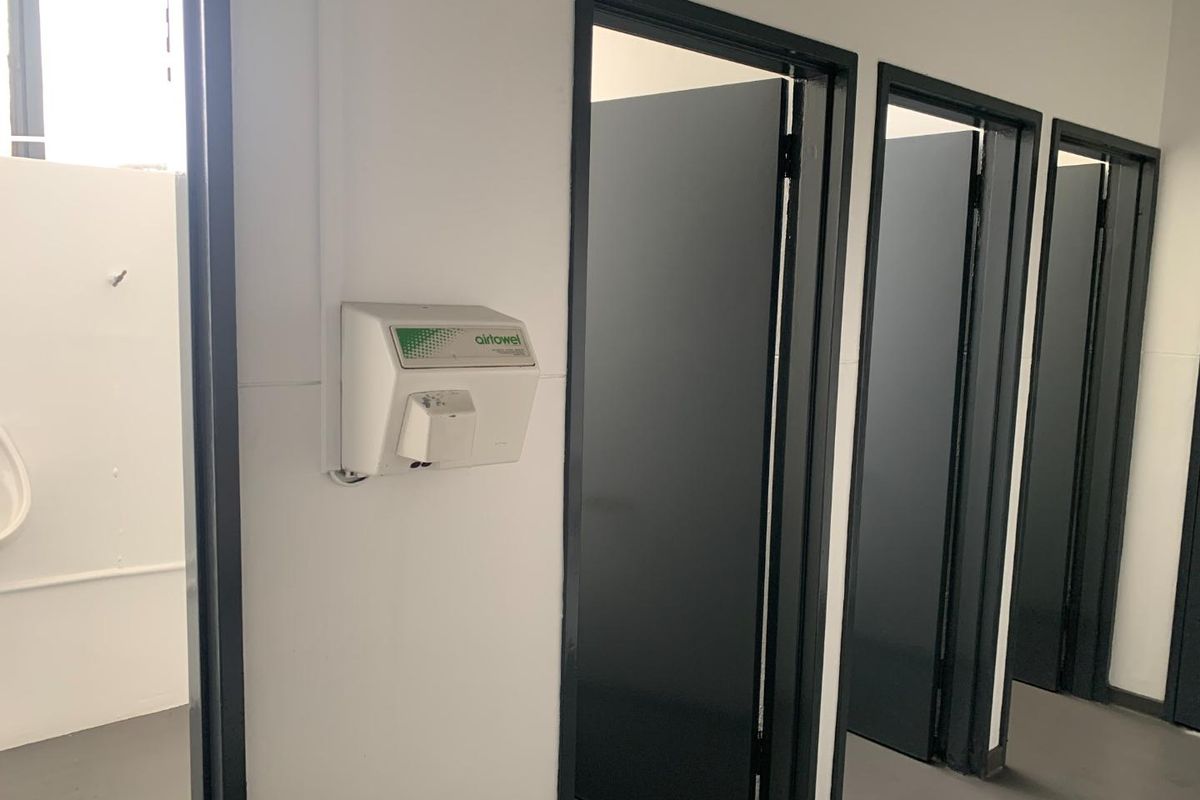 A clean industrial-style bathroom facility featuring multiple cubicle doors, a wall-mounted hand dryer, and a visible urinal.