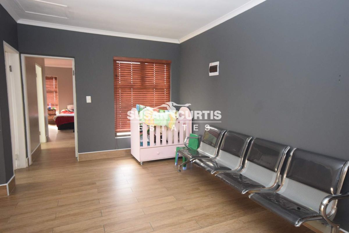 A spacious bedroom featuring wood-look tile flooring, dark grey walls, and timber venetian blinds, with an adjoining room visible through the doorway.