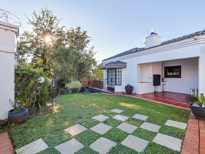 A well-maintained rear garden featuring a covered patio with a built-in braai, brick paving, and a manicured lawn area.