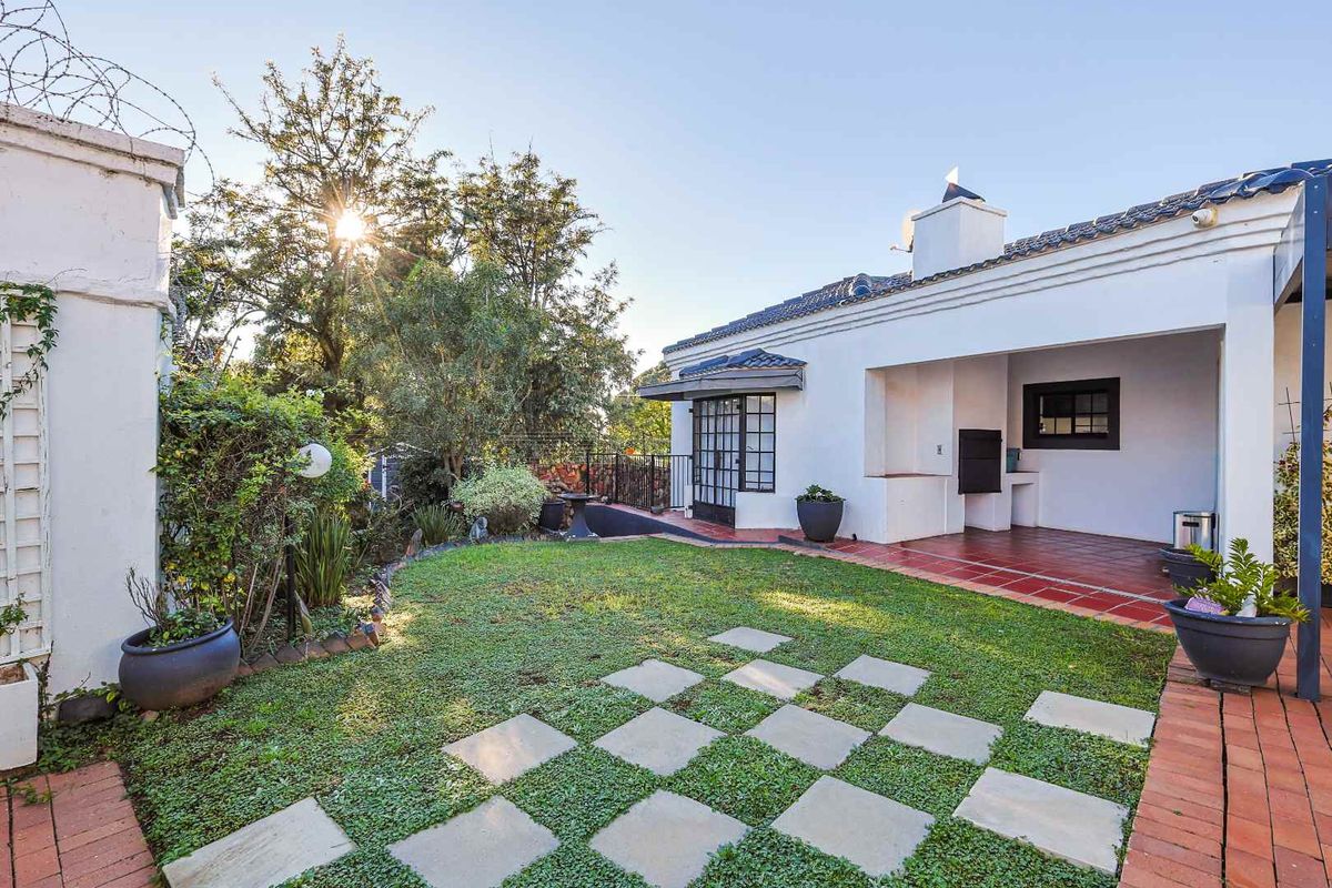 A well-maintained rear garden featuring a covered patio with a built-in braai, brick paving, and a manicured lawn area.