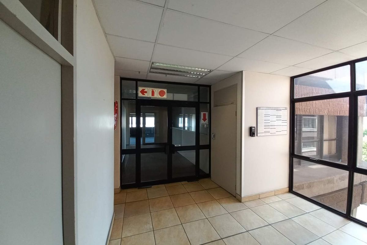 A bright office building hallway featuring tiled flooring, a suspended ceiling with recessed lighting, and large glass partitions leading to adjacent office spaces.