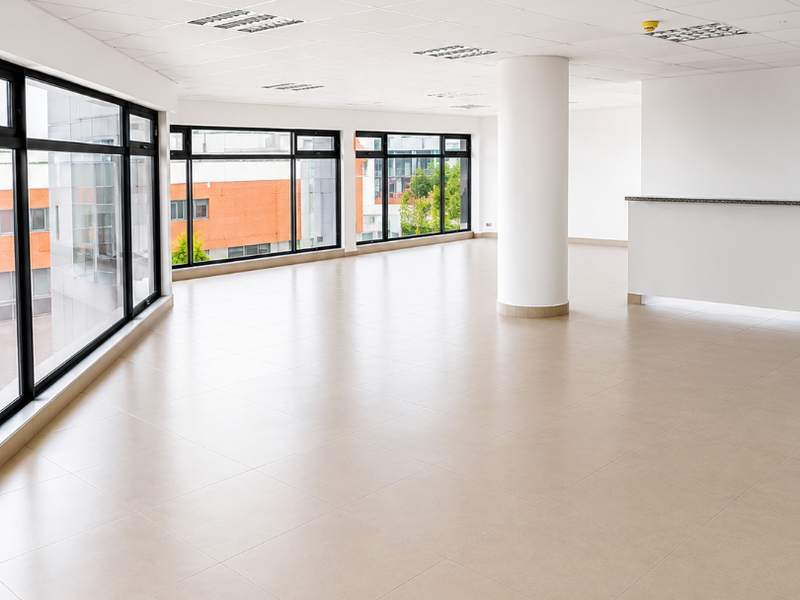 A bright, open-plan office space featuring large windows, neutral ceramic tile flooring, a suspended ceiling with recessed lighting, and a built-in reception counter.
