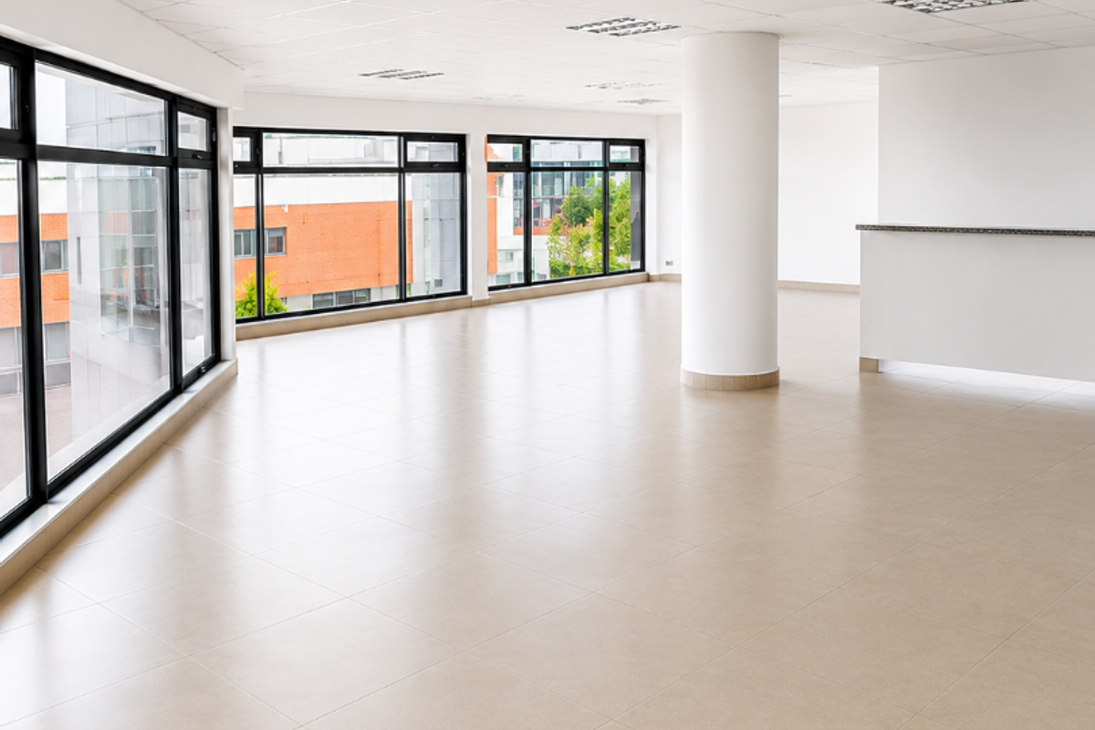 A bright, open-plan office space featuring large windows, neutral ceramic tile flooring, a suspended ceiling with recessed lighting, and a built-in reception counter.
