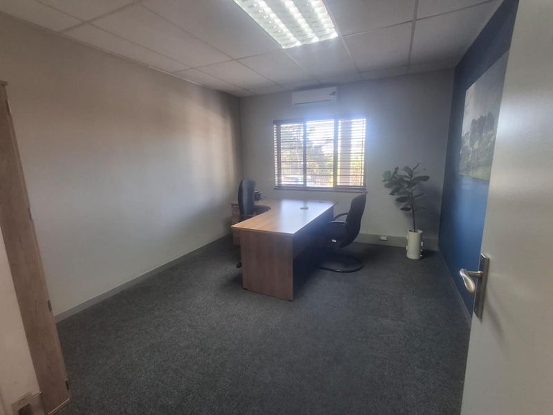 A functional office space featuring carpeted flooring, a suspended ceiling with integrated lighting, and a wall-mounted split-system air conditioner.