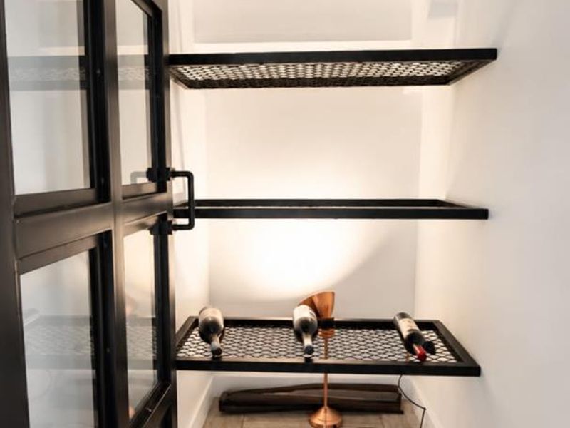 A modern, compact wine storage area featuring a sleek black metal framed glass door, built-in mesh shelving, and stylish herringbone tile flooring.
