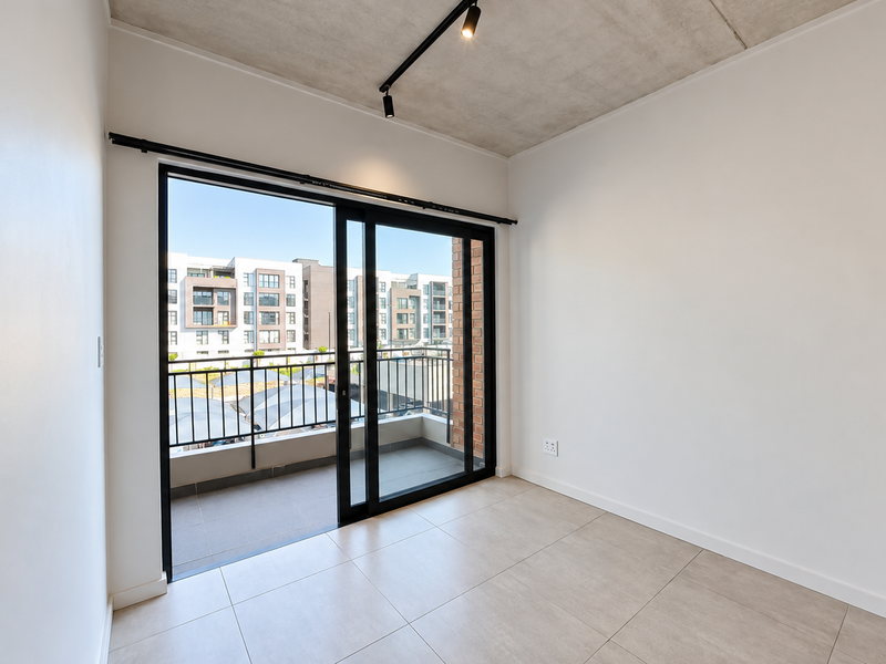 A bright, modern bedroom featuring an exposed concrete ceiling, sleek track lighting, tiled floors, and a sliding glass door leading to a private balcony.