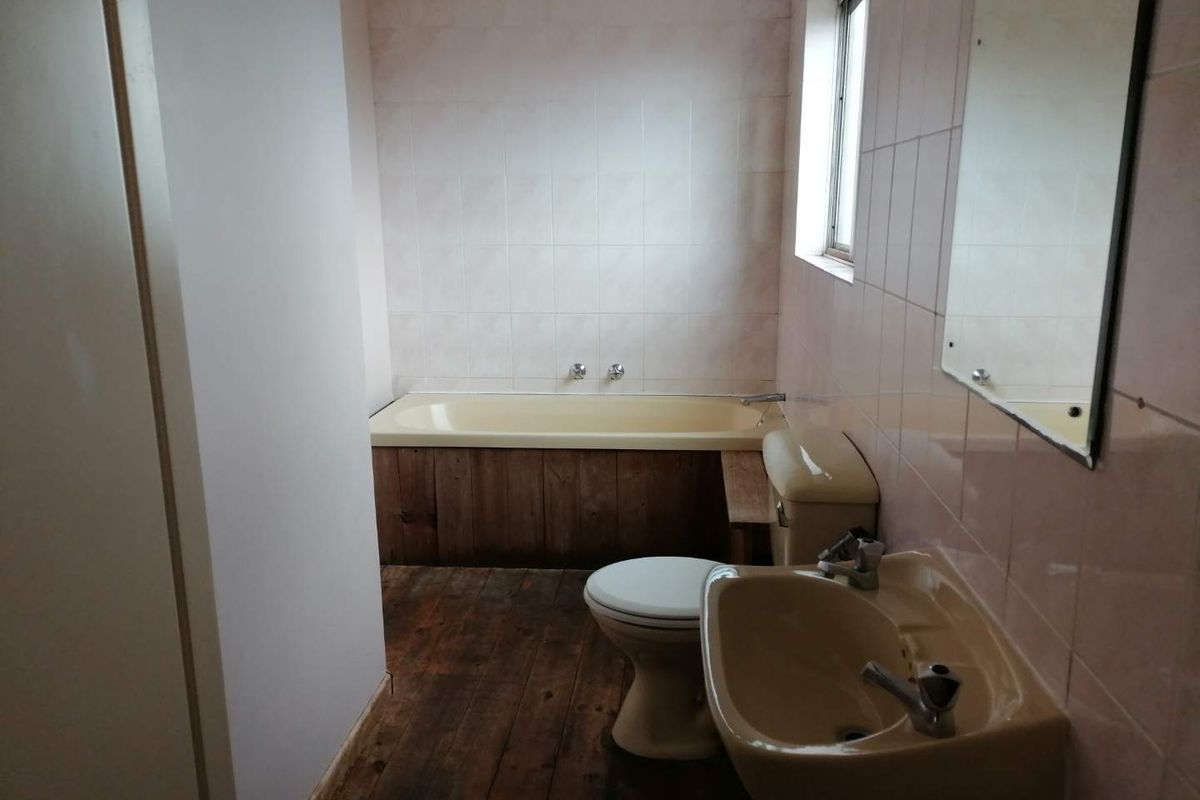 A dated bathroom featuring a vintage-style bathtub, pedestal basin, and toilet, with timber flooring and wall tiling in need of modernization.