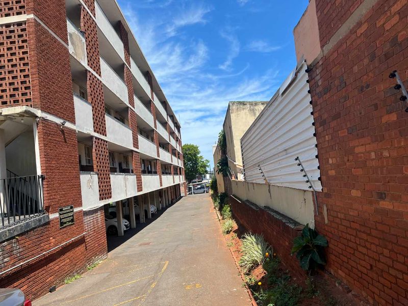 Exterior view of an apartment complex showing the rear driveway, covered parking area, and brick building facade with visible weathering.