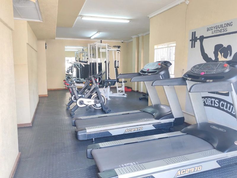 A functional gym space featuring rubber flooring, fluorescent lighting, and security bars on the windows, equipped with various fitness machines.