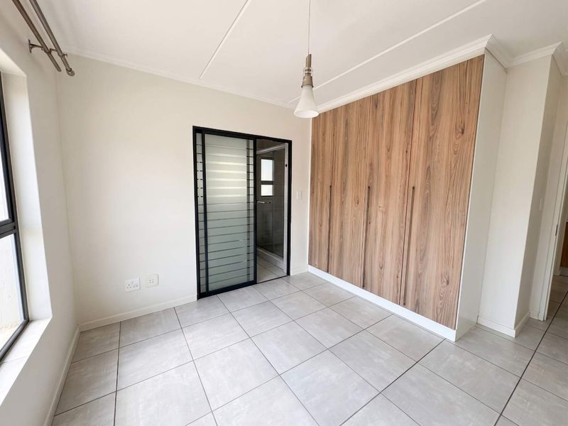 A bright, empty bedroom featuring modern wood-finish built-in cupboards, neutral tiled flooring, and a sleek sliding door leading to an en-suite bathroom.