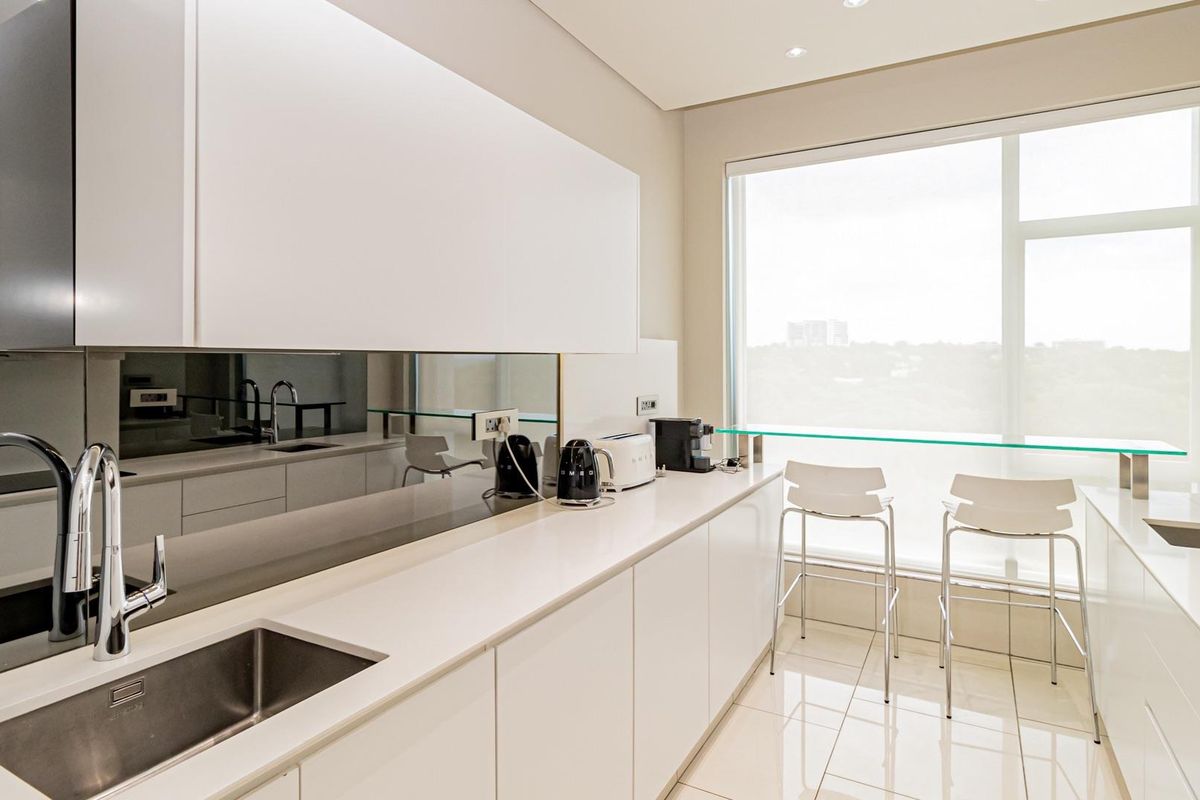 Modern galley-style kitchen featuring sleek white cabinetry, stone countertops, a mirrored backsplash, and a built-in glass breakfast bar overlooking the city view.