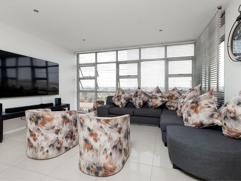 A bright and modern living room featuring large aluminium-framed windows with venetian blinds, tiled flooring, and recessed downlighting.