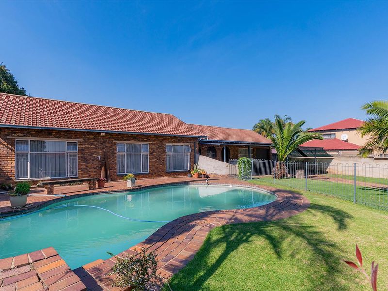 A spacious backyard featuring a swimming pool with brick paving, a well-maintained lawn, and a brick-faced house with a tiled roof.