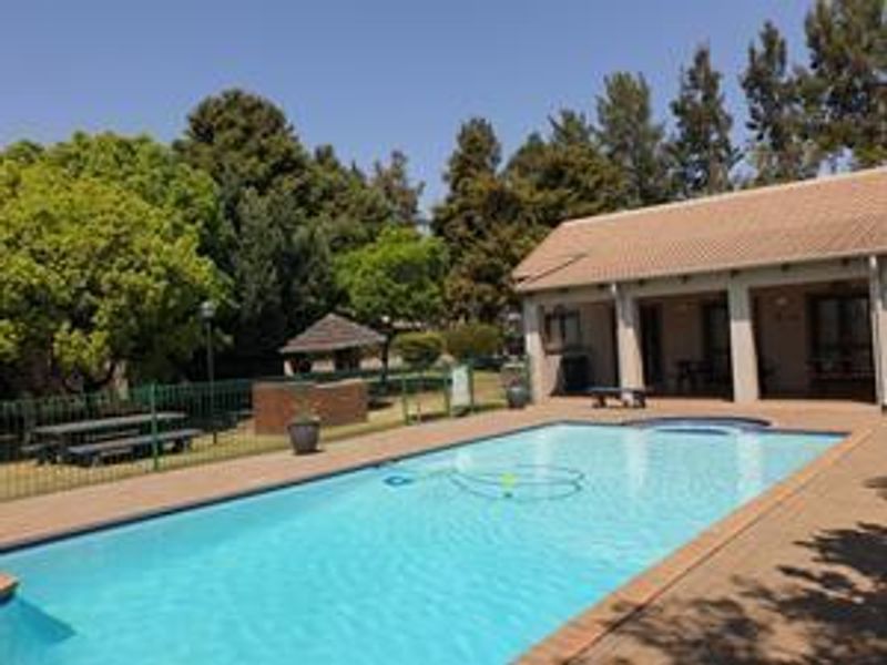 Outdoor pool area featuring a swimming pool, paved deck, brick braai facility, and a covered patio area within a landscaped garden setting.