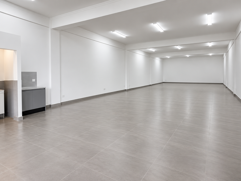 A spacious, modern open-plan office area featuring neutral tiled flooring, clean white walls, recessed lighting, and a compact built-in kitchenette.