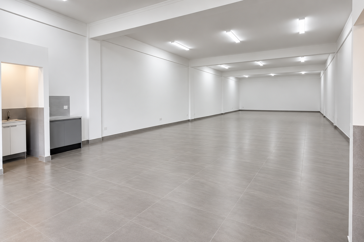 A spacious, modern open-plan office area featuring neutral tiled flooring, clean white walls, recessed lighting, and a compact built-in kitchenette.