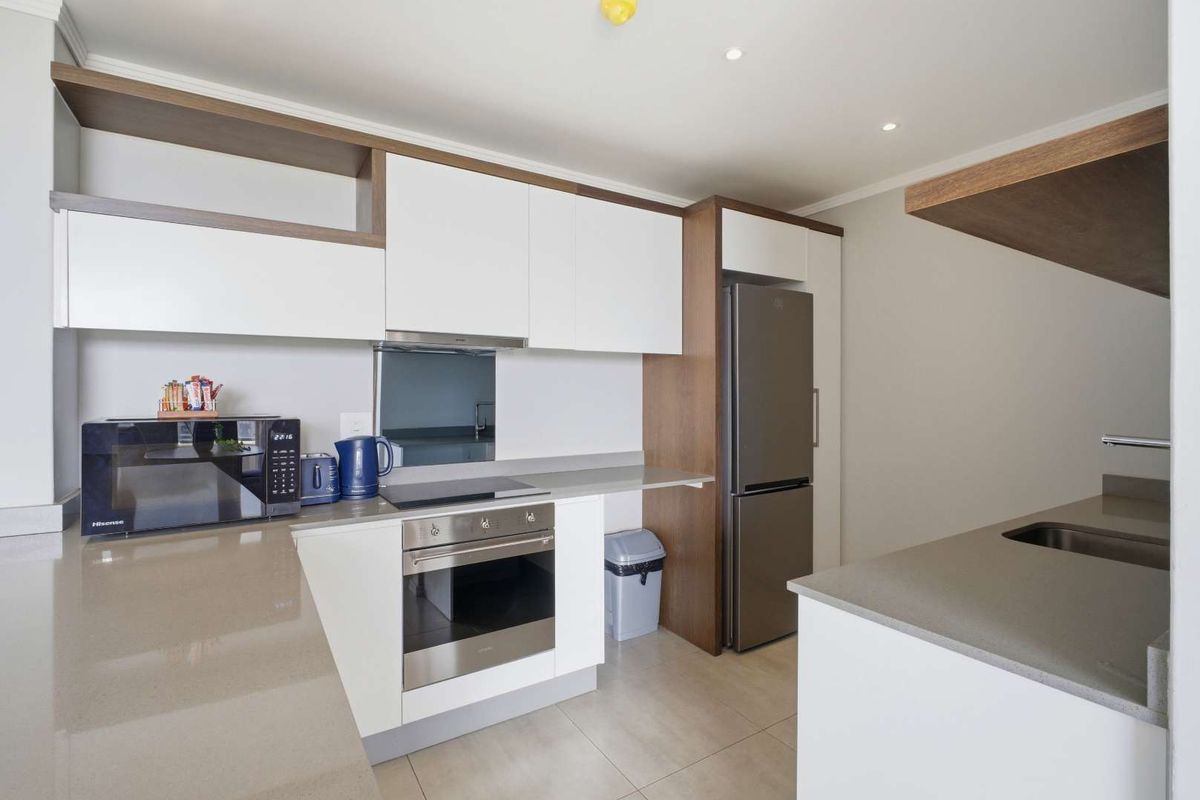 Modern kitchen featuring sleek white cabinetry, wood-tone accents, stone countertops, integrated oven, and a stainless steel sink.