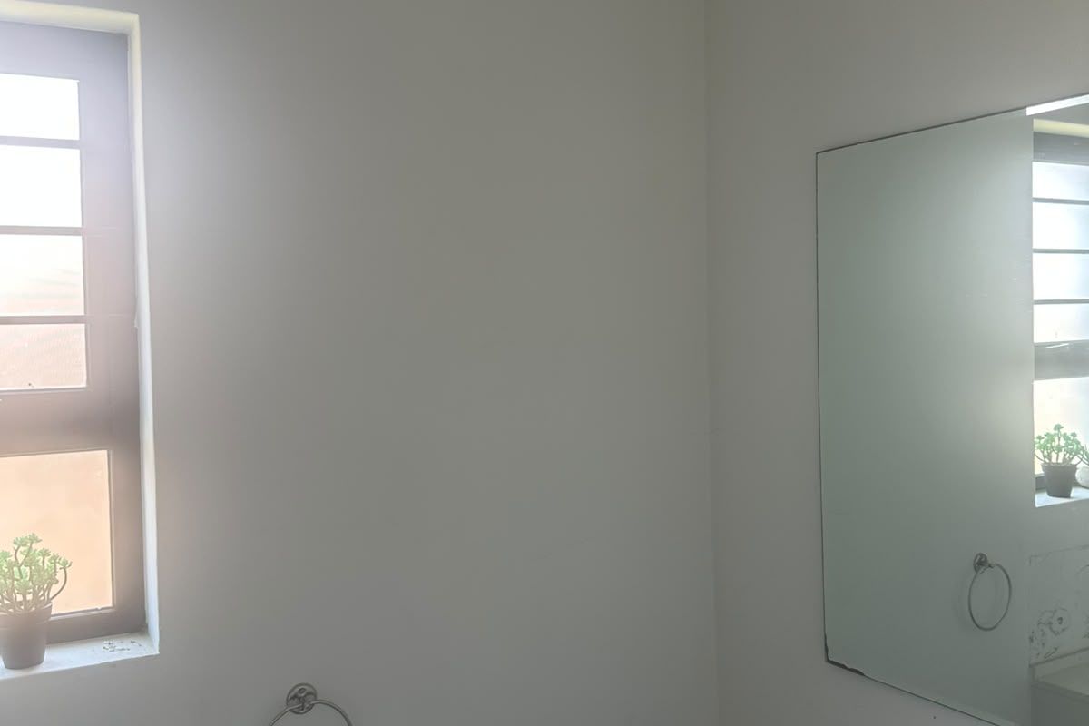 A simple bathroom interior featuring a wall-mounted mirror, towel ring, and toilet paper holder, with a window providing natural light.