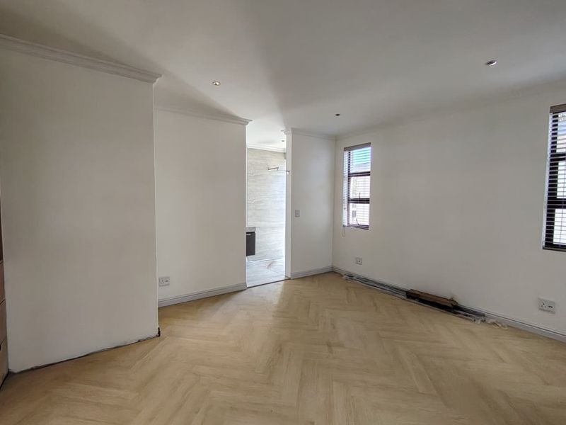 A bright, empty bedroom featuring modern herringbone flooring, recessed lighting, and clean white walls, with an adjoining bathroom visible through the doorway.