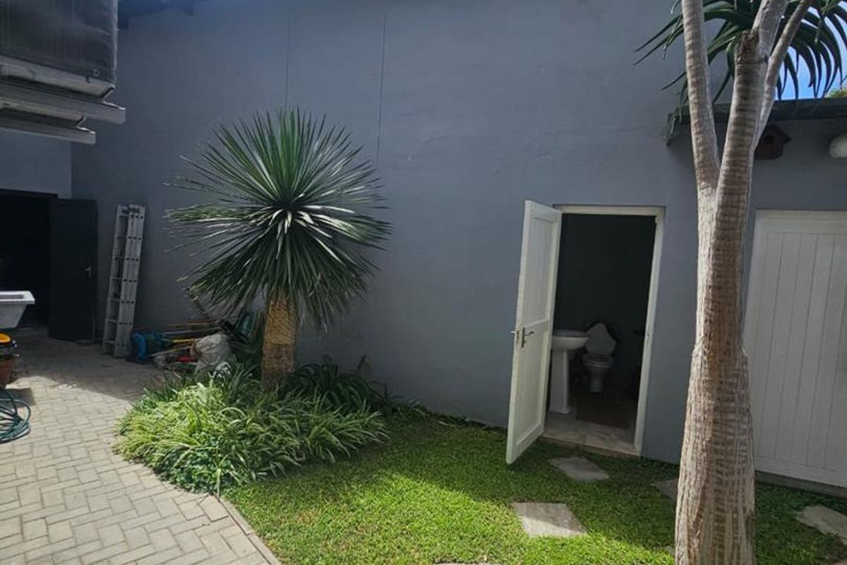 A rear exterior view featuring a paved courtyard, landscaped garden area, and access to an outdoor utility bathroom.