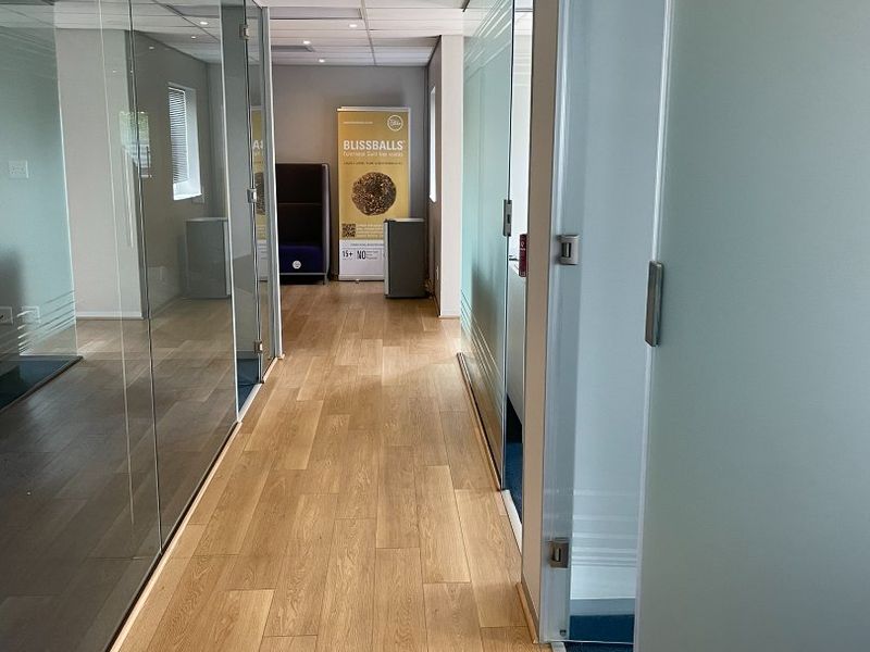 A modern office hallway featuring glass partition walls, recessed ceiling lighting, and wood-look laminate flooring.
