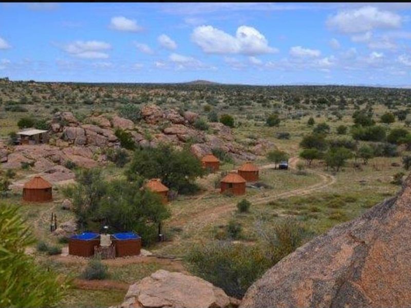 A scenic elevated view of a rural farm property featuring multiple thatched-roof accommodation units and outdoor plunge pools set within a rocky landscape.