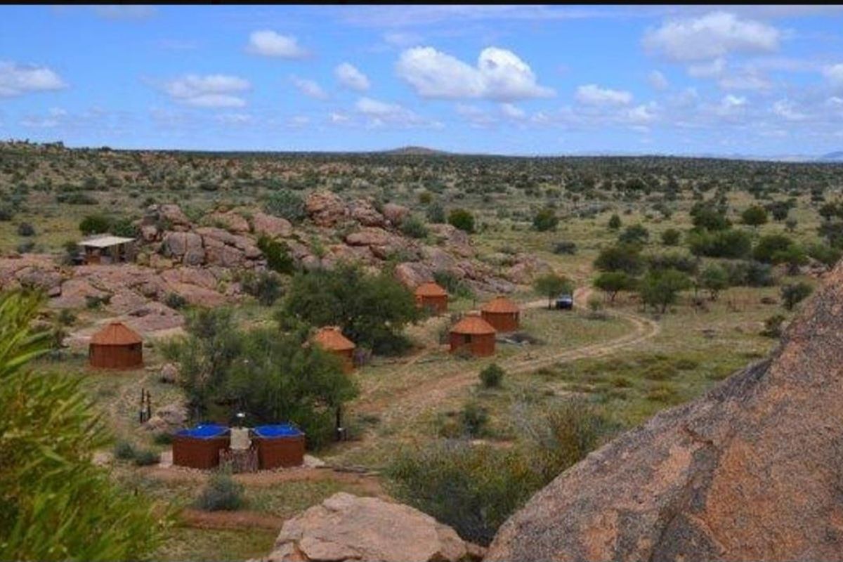 A scenic elevated view of a rural farm property featuring multiple thatched-roof accommodation units and outdoor plunge pools set within a rocky landscape.