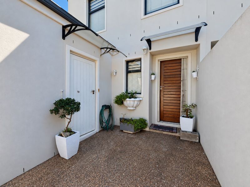 A well-maintained front entrance area featuring a timber door, secondary access door, modern awnings, and clean exposed aggregate paving.