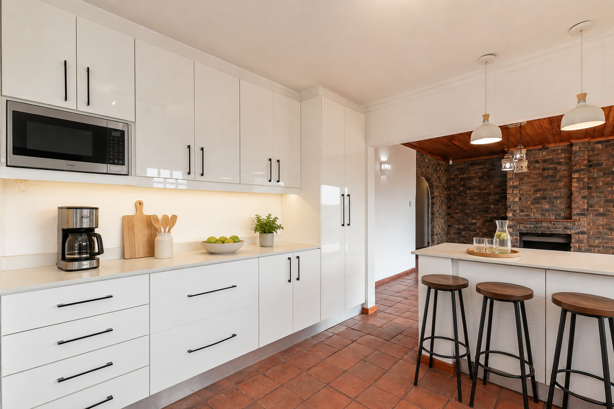 Modern kitchen featuring high-gloss white cabinetry, integrated microwave, stone countertops, and a breakfast island, complemented by rustic terracotta flooring and an exposed brick feature wall.
