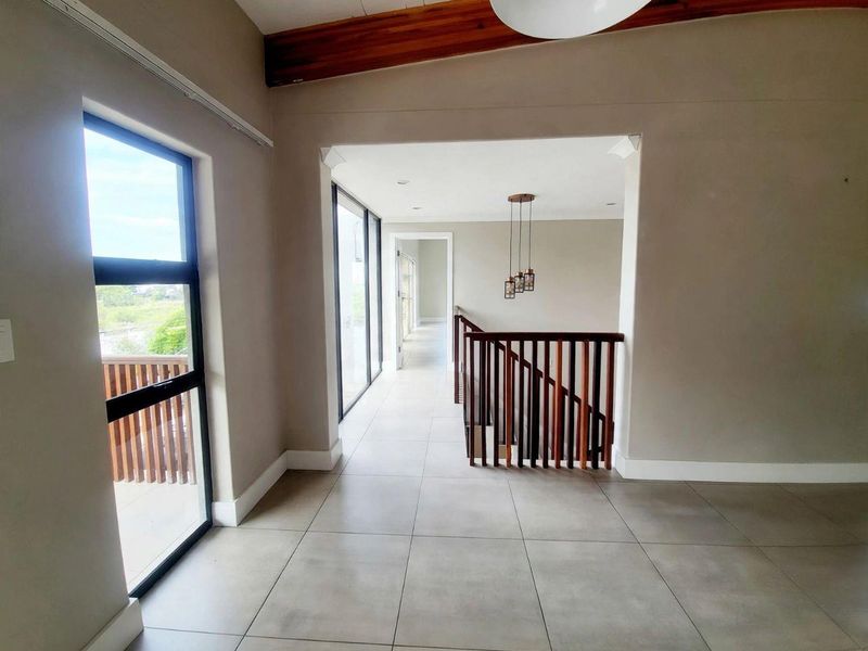 Spacious upper-level hallway featuring neutral tiled flooring, a modern wooden staircase balustrade, exposed ceiling beams, and large windows providing ample natural light.