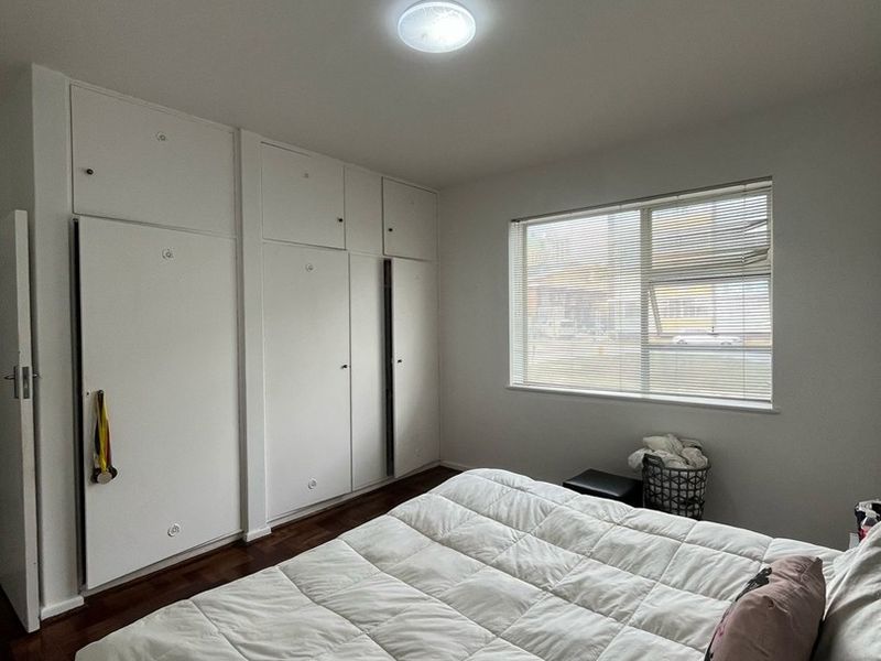 A well-maintained bedroom featuring built-in cupboards, warm parquet flooring, and a window with venetian blinds, offering a clean and functional living space.