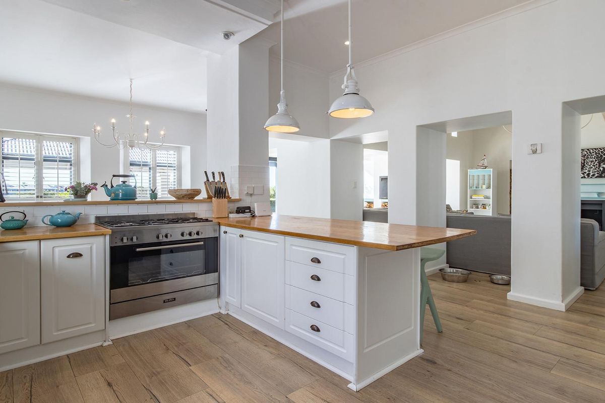 A bright, open-plan kitchen featuring white shaker-style cabinetry, timber countertops, a gas stove, and pendant lighting, seamlessly flowing into the adjacent living area.