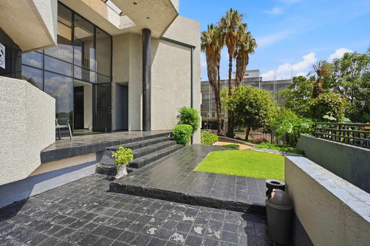 Exterior view of the office building entrance featuring a multi-level tiled patio, landscaped garden area, and modern architectural facade with large glass windows.