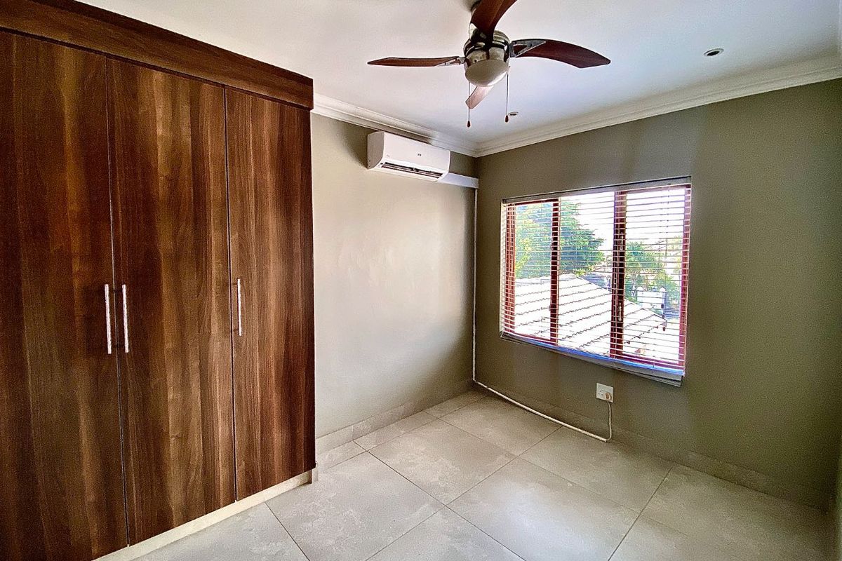 A clean, empty bedroom featuring built-in wooden cupboards, tiled flooring, a ceiling fan, and a wall-mounted air conditioning unit.