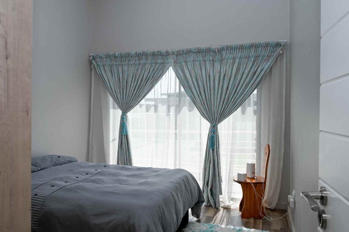 A well-lit bedroom featuring neutral wall tones, wood-look flooring, and large windows dressed with decorative curtains.