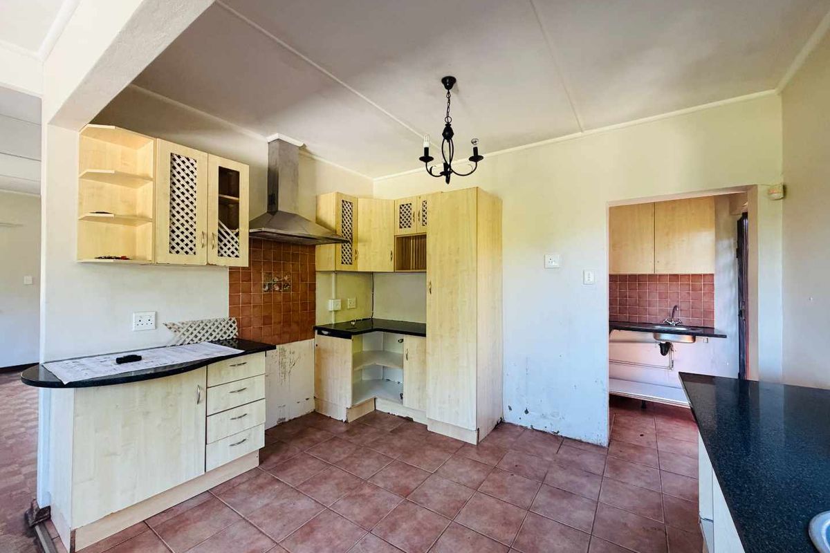 A dated kitchen featuring light wood cabinetry, dark countertops, a stainless steel extractor fan, and tiled flooring, with an adjacent scullery area.
