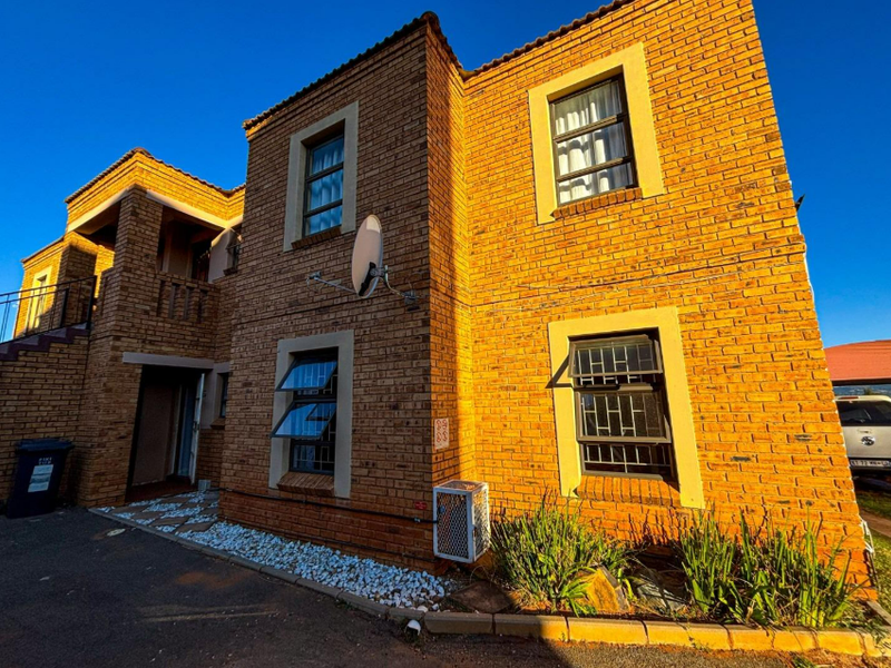A two-story face-brick townhouse exterior featuring top-hung windows with security bars, a paved driveway, and an external air conditioning unit.
